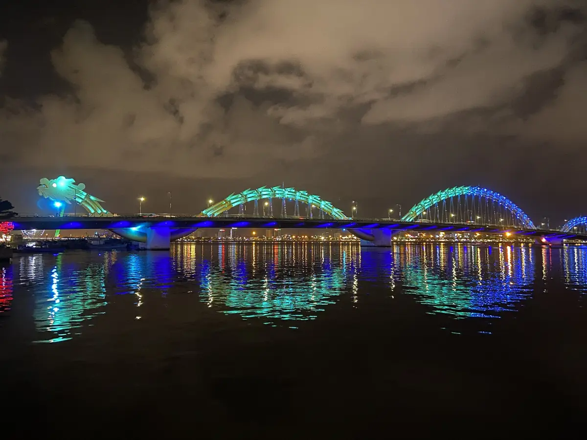 Dragon bridge lit in blue and green at night, reflected in the river